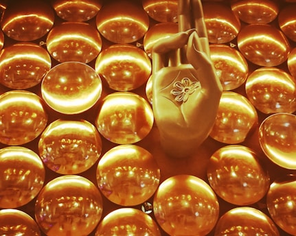 A golden hand positioned in a gesture commonly associated with meditation, surrounded by numerous shiny circular discs that create a textured backdrop. The discs reflect light, contributing to a luminous and harmonious visual effect.