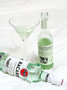 A Bacardi Carta Blanca rum bottle is lying on a white surface next to a full martini glass containing a light green liquid. A glass bottle labeled 'Cucumis' with a pale green drink inside stands upright beside them.