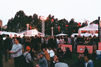 A lively outdoor event with a crowd of people gathered on grassy grounds. Some are sitting while others stand around. There are string lights and red lanterns hanging in the background, along with white tents and trees visible. The atmosphere suggests a festival or fair, possibly in the evening as lights are prominent.