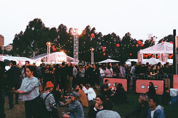 A lively outdoor event with a crowd of people gathered on grassy grounds. Some are sitting while others stand around. There are string lights and red lanterns hanging in the background, along with white tents and trees visible. The atmosphere suggests a festival or fair, possibly in the evening as lights are prominent.