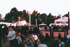 A lively outdoor event with a crowd of people gathered on grassy grounds. Some are sitting while others stand around. There are string lights and red lanterns hanging in the background, along with white tents and trees visible. The atmosphere suggests a festival or fair, possibly in the evening as lights are prominent.