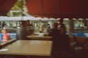 The image showcases a blurred view of people and objects in what appears to be a bus station or terminal. There are several people in motion, both inside and outside of a bus. The bus has visible text on it, and there are also tables and seats in the foreground that hint at a waiting area, possibly a cafe or rest spot.