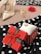 A red gift box with a bow and heart patterns sits on a polka dot fabric. Nearby is a gold ring displayed on a white cloth. In the blurred background are a pair of high-heeled shoes and a red rose, alongside some gold jewelry.