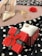 A red gift box with a bow and heart patterns sits on a polka dot fabric. Nearby is a gold ring displayed on a white cloth. In the blurred background are a pair of high-heeled shoes and a red rose, alongside some gold jewelry.