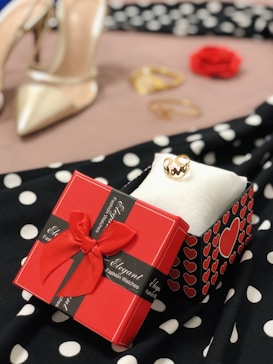 A red gift box with a bow and heart patterns sits on a polka dot fabric. Nearby is a gold ring displayed on a white cloth. In the blurred background are a pair of high-heeled shoes and a red rose, alongside some gold jewelry.