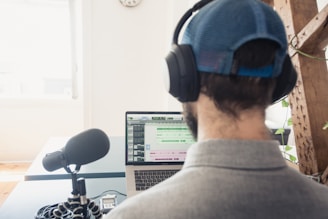 A person wearing a cap and headphones is sitting at a desk, working on a laptop. A large microphone is positioned on the desk in front of the person, suggesting audio recording or podcasting. The laptop screen displays audio editing software. There is a bright window in the background and some greenery in the room.