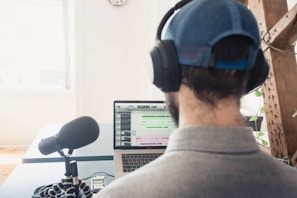 A person wearing a cap and headphones is sitting at a desk, working on a laptop. A large microphone is positioned on the desk in front of the person, suggesting audio recording or podcasting. The laptop screen displays audio editing software. There is a bright window in the background and some greenery in the room.
