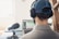 A person wearing a cap and headphones is sitting at a desk, working on a laptop. A large microphone is positioned on the desk in front of the person, suggesting audio recording or podcasting. The laptop screen displays audio editing software. There is a bright window in the background and some greenery in the room.
