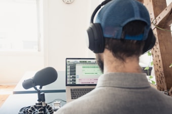 A person wearing a cap and headphones is sitting at a desk, working on a laptop. A large microphone is positioned on the desk in front of the person, suggesting audio recording or podcasting. The laptop screen displays audio editing software. There is a bright window in the background and some greenery in the room.
