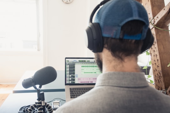 A person wearing a cap and headphones is sitting at a desk, working on a laptop. A large microphone is positioned on the desk in front of the person, suggesting audio recording or podcasting. The laptop screen displays audio editing software. There is a bright window in the background and some greenery in the room.