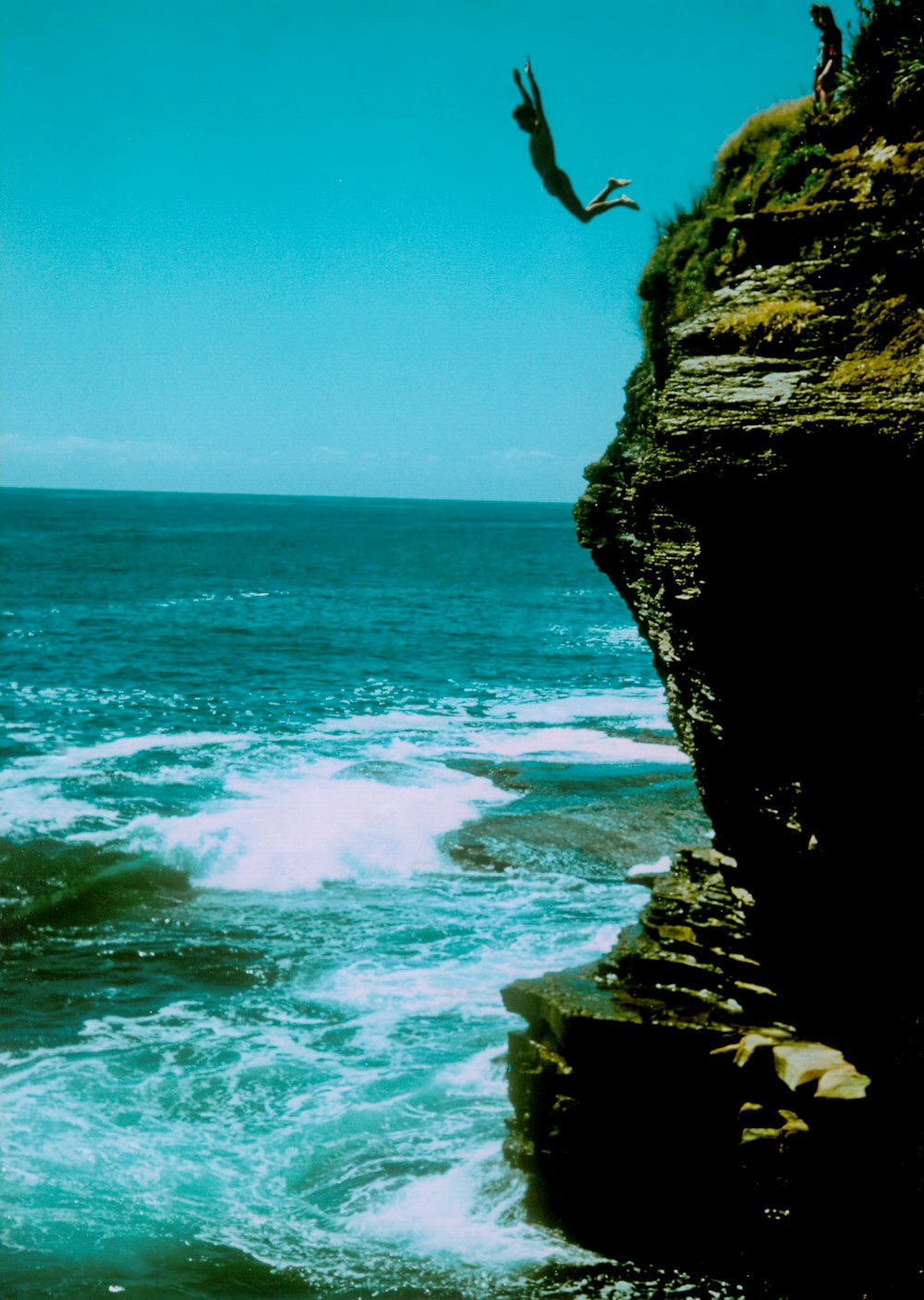 person jumping on rocky cliff near sea during daytime