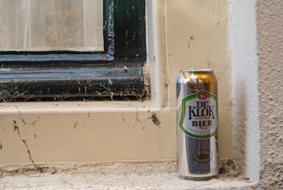 A can of De Klok beer is placed on a dusty windowsill. The window frame has a dark green wooden border, and the wall around the window is textured and pale. Cobwebs and dirt are visible around the window area, contributing to an unclean and neglected appearance.