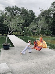 A person dressed in a white outfit and a straw hat performs a dynamic dance pose on a concrete walkway next to a pond. The person holds a colorful fabric that billows in the air, creating a striking visual contrast against the green background of lush trees and water.