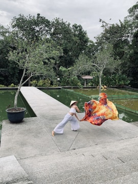 A person dressed in a white outfit and a straw hat performs a dynamic dance pose on a concrete walkway next to a pond. The person holds a colorful fabric that billows in the air, creating a striking visual contrast against the green background of lush trees and water.