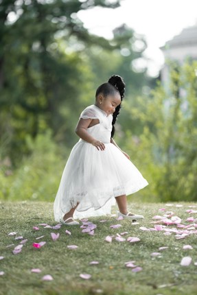 A child wearing a white dress walks gracefully on a grassy lawn adorned with scattered pink flower petals. The lush green background provides a serene and natural setting.