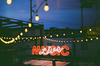 A neon sign that spells 'MUSIC' is illuminated, casting a warm glow. The scene is set at dusk or night with bokeh string lights hanging above, and blurred industrial buildings in the background.