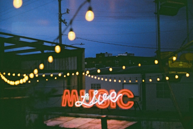 A neon sign that spells 'MUSIC' is illuminated, casting a warm glow. The scene is set at dusk or night with bokeh string lights hanging above, and blurred industrial buildings in the background.