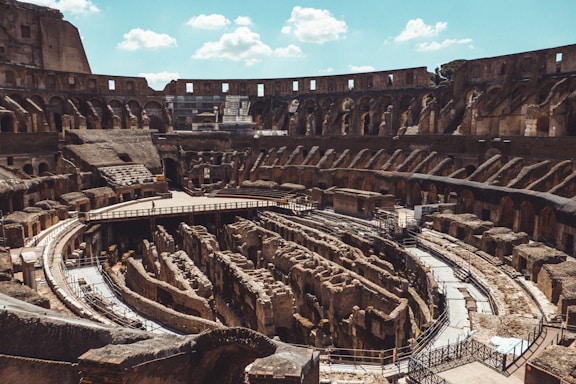 A view of the ancient Roman Colosseum showcasing its intricate architecture. The image captures the elliptical structure with its multiple levels of arches and the central arena. The exposed underground passages, which were once covered, are clearly visible. The stone and brickwork exude a sense of historical grandeur.