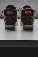 A pair of sneakers is positioned with the backs facing forward, showcasing a brown and black color scheme with the brand logo and name in red on the heel.