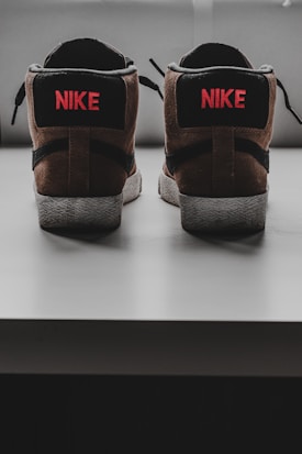 A pair of sneakers is positioned with the backs facing forward, showcasing a brown and black color scheme with the brand logo and name in red on the heel.