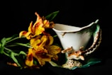 A unique ceramic cup featuring a giraffe design is placed next to a bouquet of vibrant, orange and yellow lilies with green leaves. The cup has an intricate handle resembling a giraffe's neck and head, adding artistic flair. The background is dark, highlighting the vivid colors and the intricate details of both the cup and the flowers.