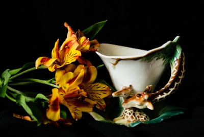 A unique ceramic cup featuring a giraffe design is placed next to a bouquet of vibrant, orange and yellow lilies with green leaves. The cup has an intricate handle resembling a giraffe's neck and head, adding artistic flair. The background is dark, highlighting the vivid colors and the intricate details of both the cup and the flowers.