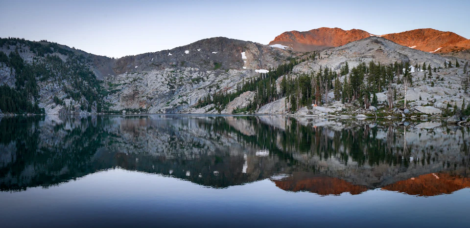 The image depicts a serene mountain landscape with a large lake at its center, surrounded by lush green trees and shrubs. The mountains are covered in snow on their peaks, creating a beautiful contrast against the clear blue sky above. The sun is setting behind the mountains, casting a warm glow over the scene and enhancing the colors of the landscape.
