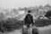 A person is walking along a path with two rolling suitcases. The person is wearing a backpack and is seen from behind, facing a view of a town or small city with numerous houses and trees in the background. The scene suggests travel or movement towards a destination.