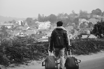 A person is walking along a path with two rolling suitcases. The person is wearing a backpack and is seen from behind, facing a view of a town or small city with numerous houses and trees in the background. The scene suggests travel or movement towards a destination.