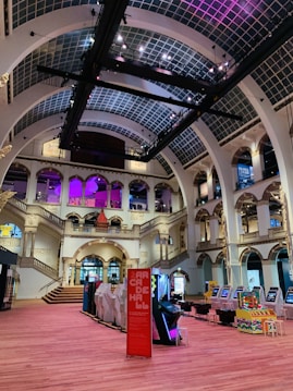 The interior of a large, ornately designed building with arched ceilings and intricate architectural details. The space features gaming machines lined up along the floor, under a high ceiling with a grid pattern. Lighting in purple and white tones enhances the ambiance.
