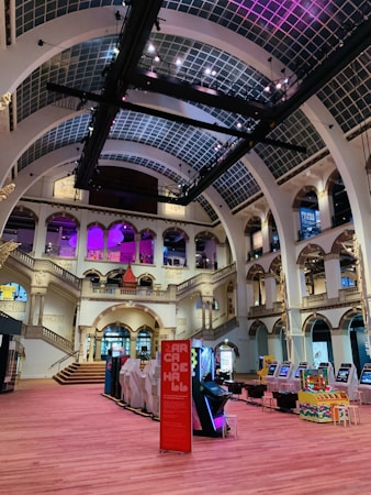 The interior of a large, ornately designed building with arched ceilings and intricate architectural details. The space features gaming machines lined up along the floor, under a high ceiling with a grid pattern. Lighting in purple and white tones enhances the ambiance.