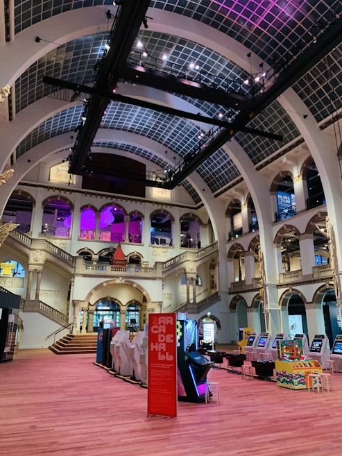 The interior of a large, ornately designed building with arched ceilings and intricate architectural details. The space features gaming machines lined up along the floor, under a high ceiling with a grid pattern. Lighting in purple and white tones enhances the ambiance.