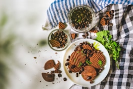 A plate of chocolate cookies garnished with chocolate chips and fresh herbs. Several cookies are scattered on a white surface with coffee beans around them. Two glass jars filled with coffee beans are placed on a folded cloth with blue and black checkered patterns. A sprig of green leaves adds a touch of freshness.