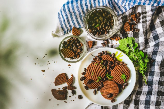 A plate of chocolate cookies garnished with chocolate chips and fresh herbs. Several cookies are scattered on a white surface with coffee beans around them. Two glass jars filled with coffee beans are placed on a folded cloth with blue and black checkered patterns. A sprig of green leaves adds a touch of freshness.