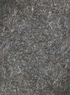 A close-up view of textured asphalt road surface with visible rough and bumpy aggregate particles. The image captures the gritty details and uneven nature of the asphalt material.