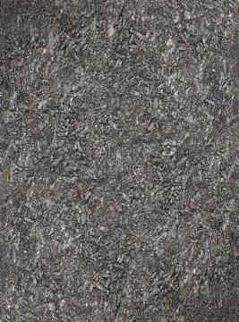 A close-up view of textured asphalt road surface with visible rough and bumpy aggregate particles. The image captures the gritty details and uneven nature of the asphalt material.