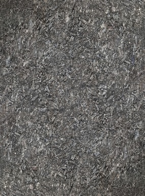 A close-up view of textured asphalt road surface with visible rough and bumpy aggregate particles. The image captures the gritty details and uneven nature of the asphalt material.