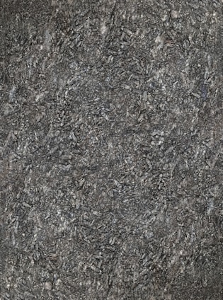 A close-up view of textured asphalt road surface with visible rough and bumpy aggregate particles. The image captures the gritty details and uneven nature of the asphalt material.