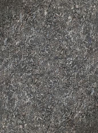 A close-up view of textured asphalt road surface with visible rough and bumpy aggregate particles. The image captures the gritty details and uneven nature of the asphalt material.