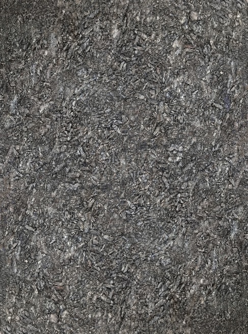 A close-up view of textured asphalt road surface with visible rough and bumpy aggregate particles. The image captures the gritty details and uneven nature of the asphalt material.