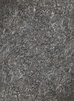 A close-up view of textured asphalt road surface with visible rough and bumpy aggregate particles. The image captures the gritty details and uneven nature of the asphalt material.