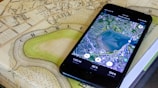 A smartphone displaying a satellite view of a large body of water is placed on an old, detailed map. The phone screen shows a map application with various measurements and geographical details. The map beneath appears to be historical, with intricate street layouts and labels.