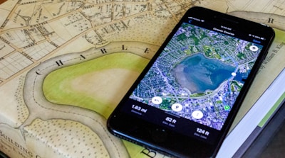 A smartphone displaying a satellite view of a large body of water is placed on an old, detailed map. The phone screen shows a map application with various measurements and geographical details. The map beneath appears to be historical, with intricate street layouts and labels.