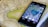 A smartphone displaying a satellite view of a large body of water is placed on an old, detailed map. The phone screen shows a map application with various measurements and geographical details. The map beneath appears to be historical, with intricate street layouts and labels.