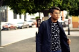 A young person is standing outdoors on a street, wearing a dark jacket over a graphic t-shirt with geometric designs. They are looking to the side, and the background features a few parked cars and buildings with trees providing shade.