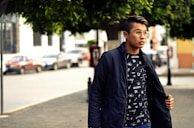 A young person is standing outdoors on a street, wearing a dark jacket over a graphic t-shirt with geometric designs. They are looking to the side, and the background features a few parked cars and buildings with trees providing shade.