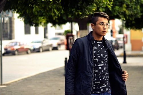 A young person is standing outdoors on a street, wearing a dark jacket over a graphic t-shirt with geometric designs. They are looking to the side, and the background features a few parked cars and buildings with trees providing shade.