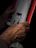 An elderly hand holding a magazine or book, with visible wrinkles and a focus on the hand's texture. The magazine has a prominent red edge and some text and an image are slightly blurred in the background.