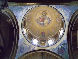 A richly decorated dome interior featuring a central religious icon surrounded by angels and other biblical figures, all portrayed in traditional Eastern Orthodox style. The colors are vibrant, with the use of blue, gold, and red. Architectural details and inscriptions enhance the spiritual atmosphere.