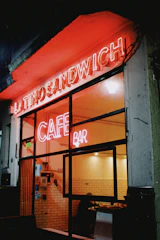 A nighttime scene features the entrance of a cafe bar with bright neon signs glowing in red that read 'LATINO SANDWICH' and 'CAFE BAR'. The interior, visible through the glass front, shows a warmly lit space with white-tiled walls and a person sitting at a table.